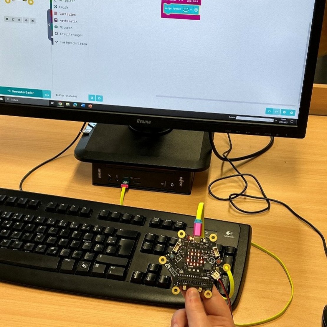 Eine Person sitzt am Tisch und hält ein kleines Gerät in der Hand. Das Gerät ist mit dem Computer verbunden. Auf dem Bildschirm sieht man bunte Kästchen, die zum Programmieren da sind.