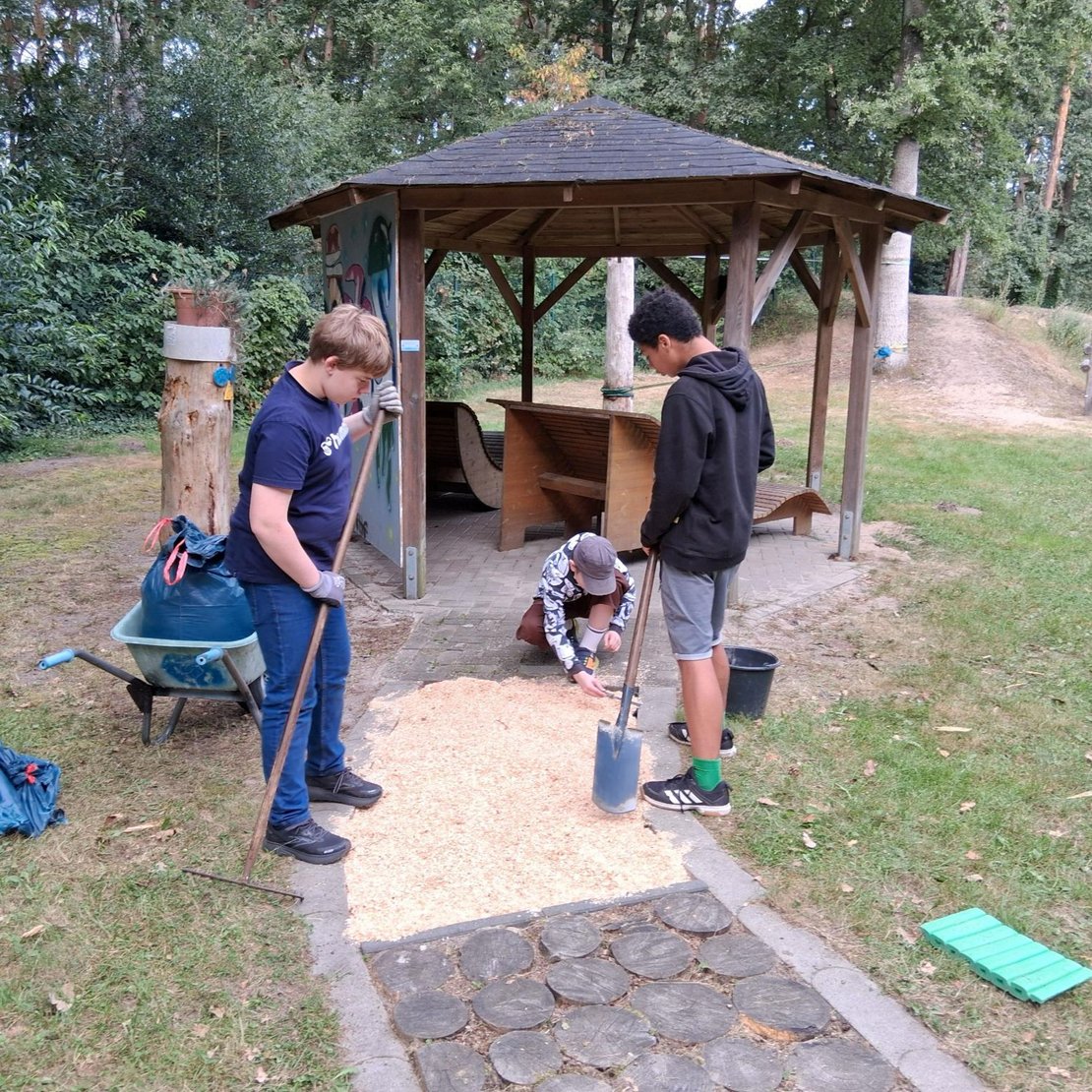 Drei Personen arbeiten gemeinsam an einem Weg neben einem hölzernen Pavillon in einer grünen Außenanlage. Eine Person steht mit einem Rechen, eine andere hält eine Schaufel, und die dritte kniet und verteilt Holzschnitzel. Rundherum liegen Werkzeuge wie eine Schubkarre, eine blaue Plane und ein grünes Kunststoffobjekt.