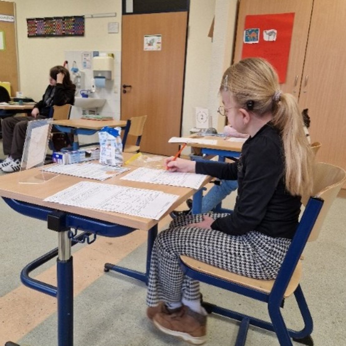 In einem Klassenraum sitzen Schüler an Tischen. Ein Schüler schreibt etwas auf ein Blatt Papier. An den Wänden hängen Bilder und Lernmaterialien.
