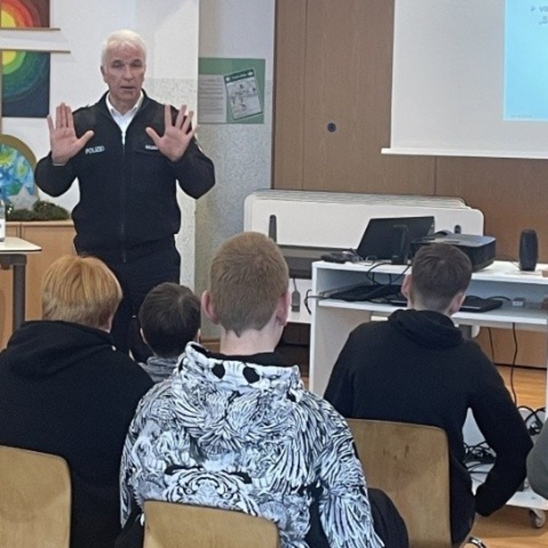 Ein Mann steht vor einer Gruppe von Schülern in einem Klassenraum. Er spricht und hat beide Hände gehoben. Die Schüler sitzen auf Holzstühlen und hören zu. Im Hintergrund ist eine Leinwand mit Text zu sehen.