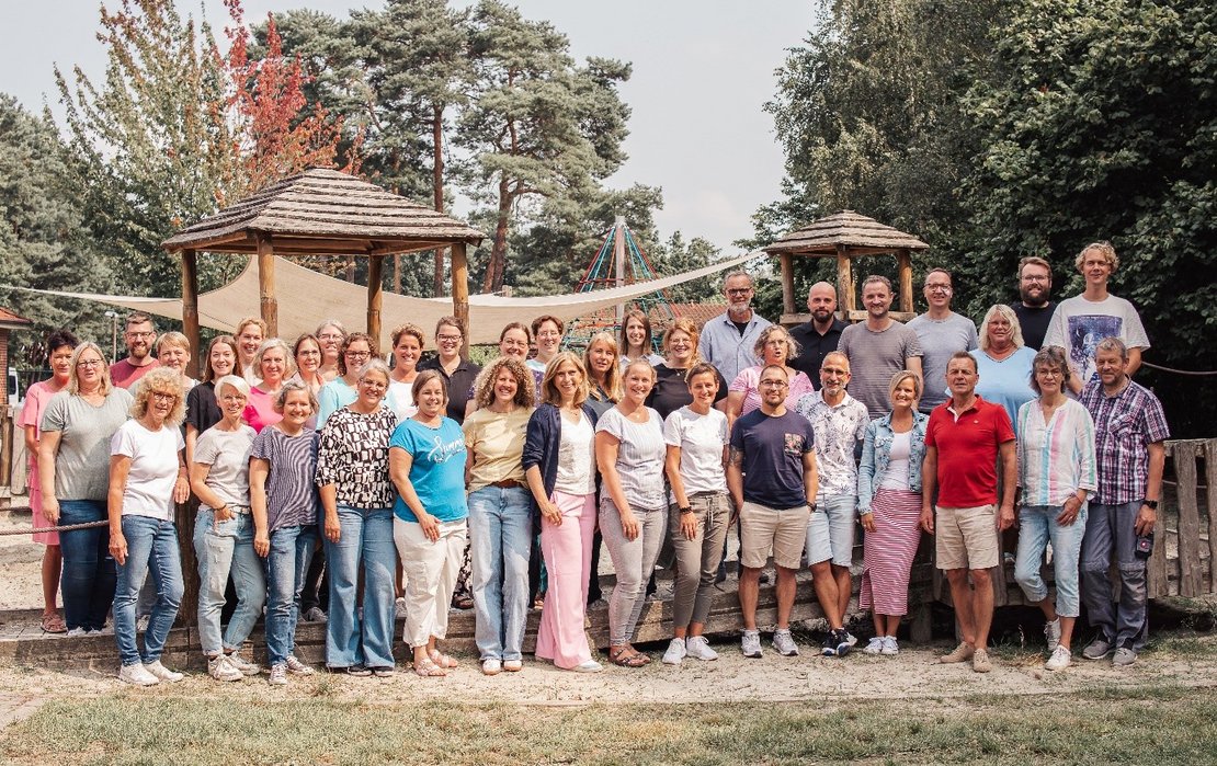 Gruppe von Menschen im Freien auf einem Spielplatz, mit Bäumen und Holzstrukturen im Hintergrund.