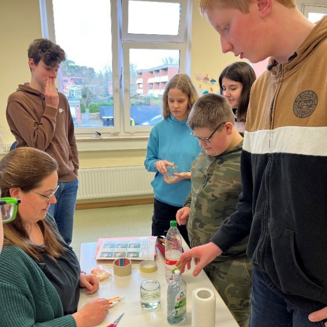 Mehrere Menschen sitzen an einem Tisch in einem Klassenraum. Auf dem Tisch liegen Alufolie, Papier, Schere, Klebeband, ein Glas mit Flüssigkeit und Spülmittel. Die Gruppe macht gemeinsam ein Experiment oder eine Bastelarbeit.