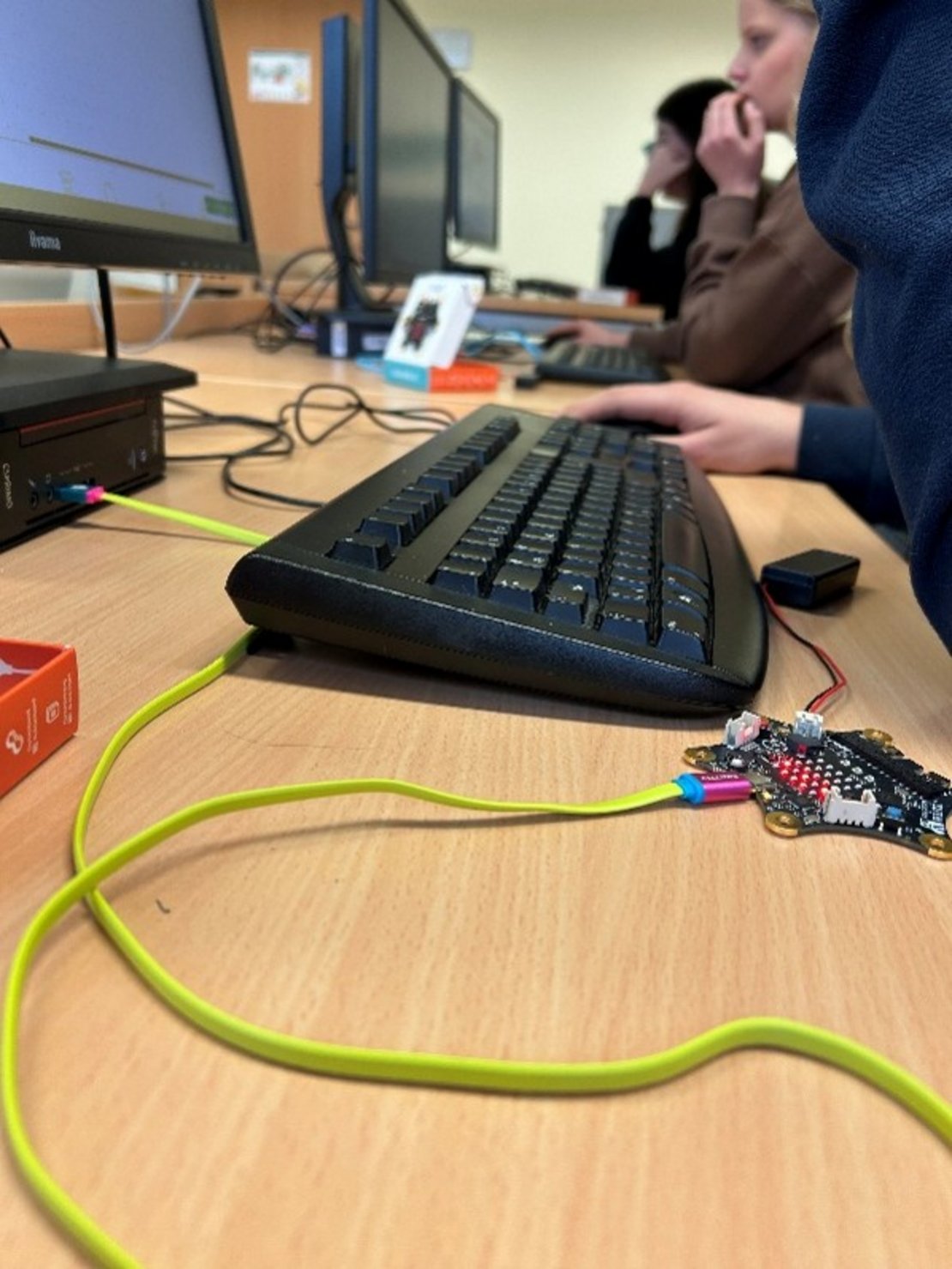 In einem Klassenraum stehen mehrere Computer auf einem Holztisch. Man sieht Tastaturen, Bildschirme und Kabel. Im Vordergrund sind grüne Kabel und elektronische Teile zu sehen. Zwei Personen arbeiten im Hintergrund an den Computern.