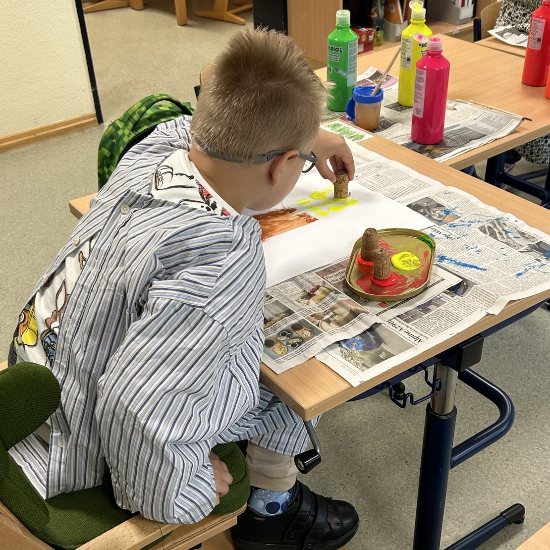 Eine Person mit Brille und gestreiftem Shirt sitzt an einem Tisch und stempelt gelbe Farbe mit einem Korken auf ein bemaltes Blatt Papier. Der Tisch ist mit Zeitungspapier abgedeckt, darauf stehen mehrere Farbtöpfe in Grün, Gelb und Pink. Die Person trägt eine Schutzkleidung gegen Farbspritzer. Es handelt sich um eine kreative Malaktivität.