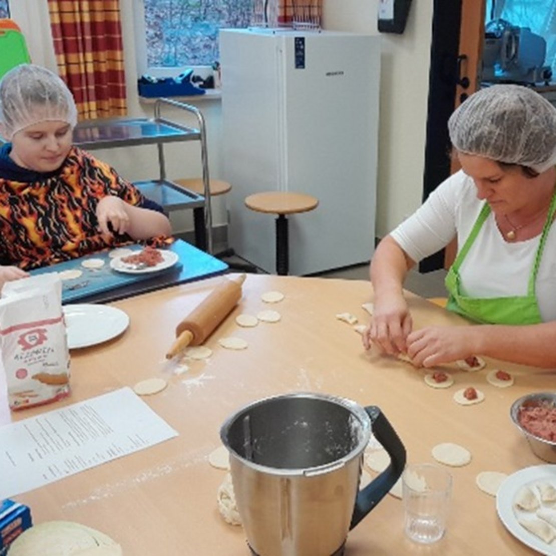Mehrere Menschen stehen in einer Küche. Sie tragen Haarnetze und Schürzen. Auf dem Tisch liegen Teig, Mehl und eine Schüssel mit Füllung. Sie machen zusammen kleine Teigtaschen.