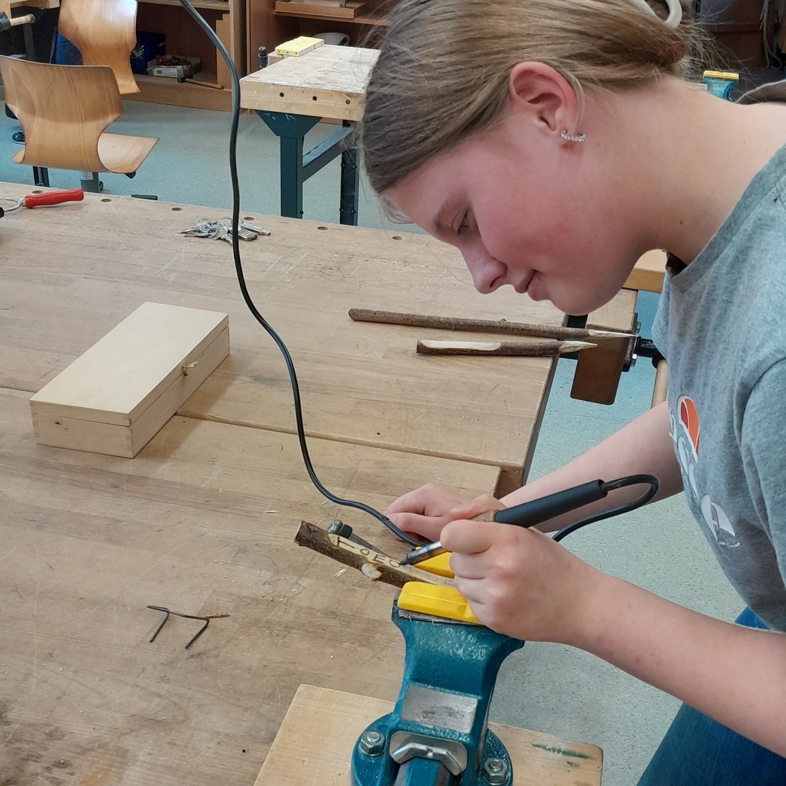 Person arbeitet in einer Werkstatt mit einem Lötkolben an einem Objekt, das in einem blauen Schraubstock auf einer Holzwerkbank eingespannt ist. Auf der Werkbank liegen Werkzeuge, Holzstücke und Metallstäbe. Im Hintergrund sind Regale mit weiteren Werkzeugen und Materialien zu sehen.